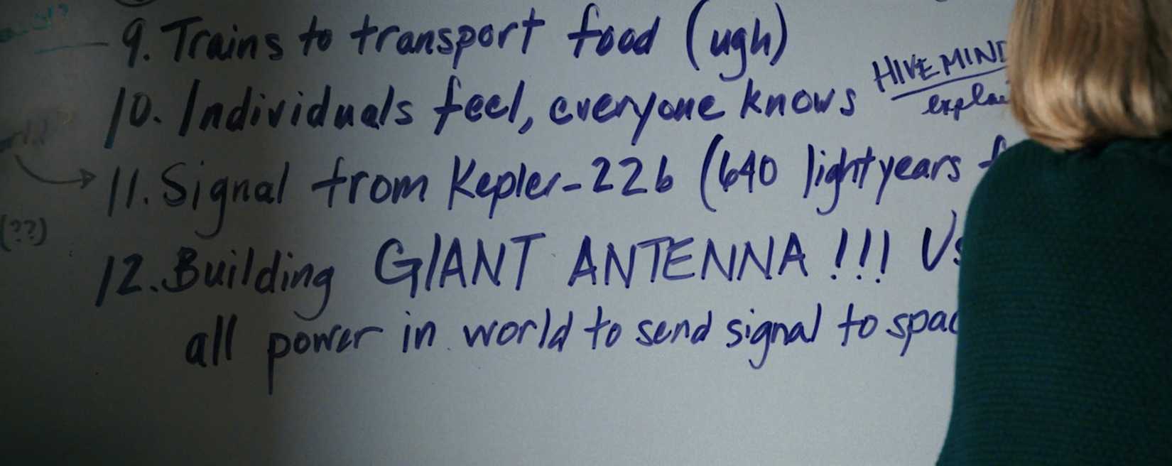 Carol (Rhea Seehorn) stands with her back to the camera, facing a whiteboard covered with facts about the Others. Prominently in the middle, in all caps, are the words "GIANT ANTENNA!!!"